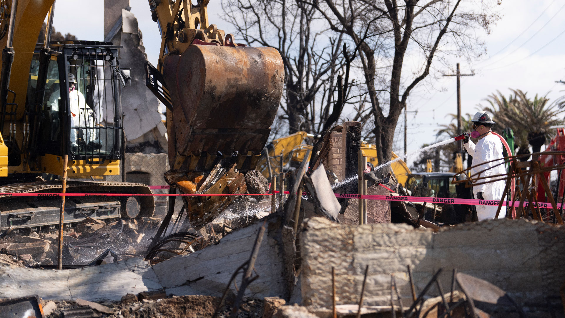 Crews contracted by the Army Corps of Engineers operate an excavator and hose to clear fire debris from a private property.