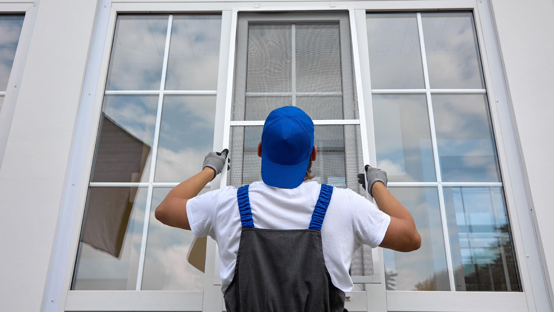 A man in a backwards blue hat installs a window pane on a home.