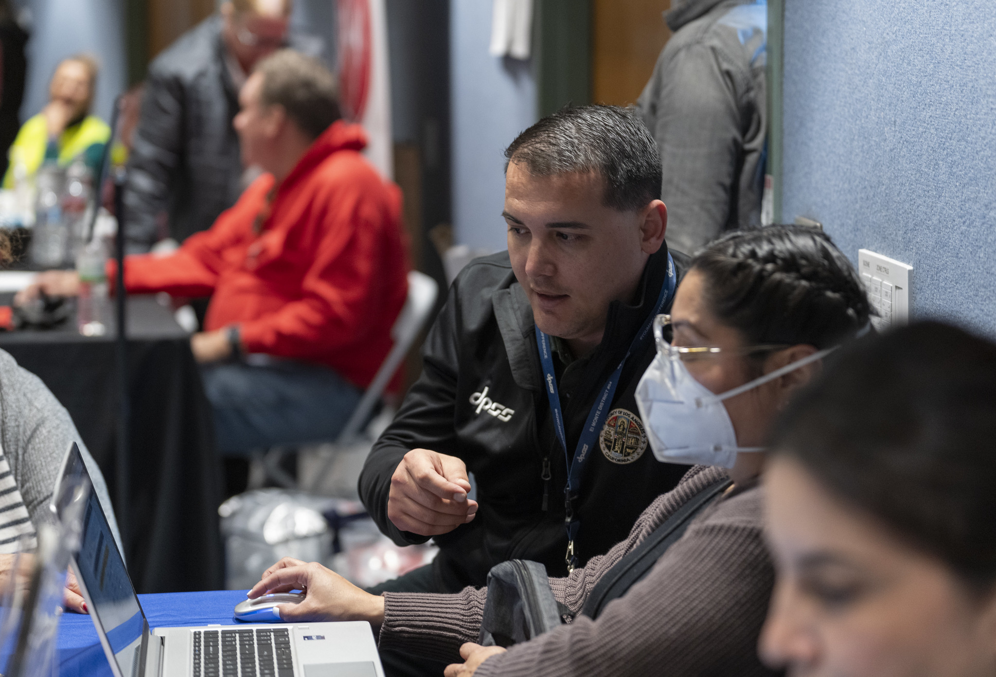 A man in a black jacket assists a woman in a mask at a table with laptops during an event.